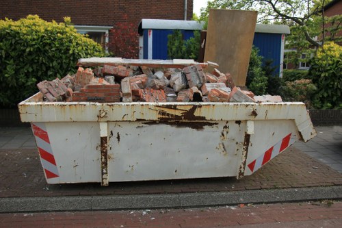A large, white skip container with red and white diagonal stripe markings on the sides, positioned on a residential street in Hainault. The skip is filled with a piled heap of irregularly shaped red bricks and concrete debris, some with visible mortar residue. The surface of the skip shows rust and stains, indicating exposure to outdoor elements. Behind the skip, a paved pavement runs alongside a brick wall, with a neatly trimmed hedge and a blue wooden shed visible in the background. A large wooden panel leans against the shed, adding to the clutter of construction debris within the skip. The scene is captured during daylight, with ambient natural light illuminating the area, emphasizing the textures and colors of the brick and concrete refuse consistent with garden clearance and rubbish removal services in the Hainault area.