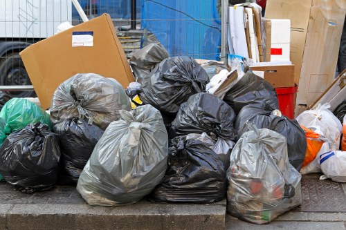 A collection of black and grey plastic garbage bags filled with mixed waste, placed on a paved area, possibly a driveway or pavement in Hainault. The bags are tightly knotted and stacked in front of cardboard boxes and other miscellaneous rubbish materials, some of which are partially visible behind the bags. The background includes a stack of white and brown boxes, a red container, and a blue wooden structure, suggesting an outdoor storage or waste collection point. The scene appears to be part of a rubbish removal project or garden clearance in the local area, with the bags organized for collection or disposal by Garden Clearance Hainault. The environment is well-lit, with natural daylight illuminating the scene, emphasizing the different textures of the plastic, cardboard, and surrounding objects, and indicating an active effort to clear accumulated waste in a residential or commercial space in Hainault or nearby postcode areas. This detailed visual context aligns with professional rubbish removal services provided in the local community.