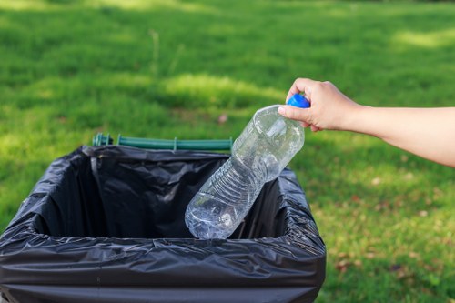 A person’s hand is seen placing a clear plastic water bottle into a black rubbish bin liner that is lining a partially visible black wheeled bin or trash container. The water bottle appears to be a typical PET plastic container with a blue screw cap, and it is being dropped in from the right side of the image. The background showcases a well-maintained, lush green lawn with blurred grass, suggesting an outdoor setting such as a garden or park area. The scene is well-lit with natural daylight, highlighting the textures of the plastic bottle and the smooth surface of the black rubbish bin liner. The positioning indicates an act of rubbish disposal, possibly as part of garden waste or general waste removal services provided by Garden Clearance Hainault, located in the Hainault area near the postcode corridor. The overall context aligns with professional waste management activities involving household or garden refuse collection based in local Essex or Greater London surroundings, emphasizing environmental responsibility and cleanliness.