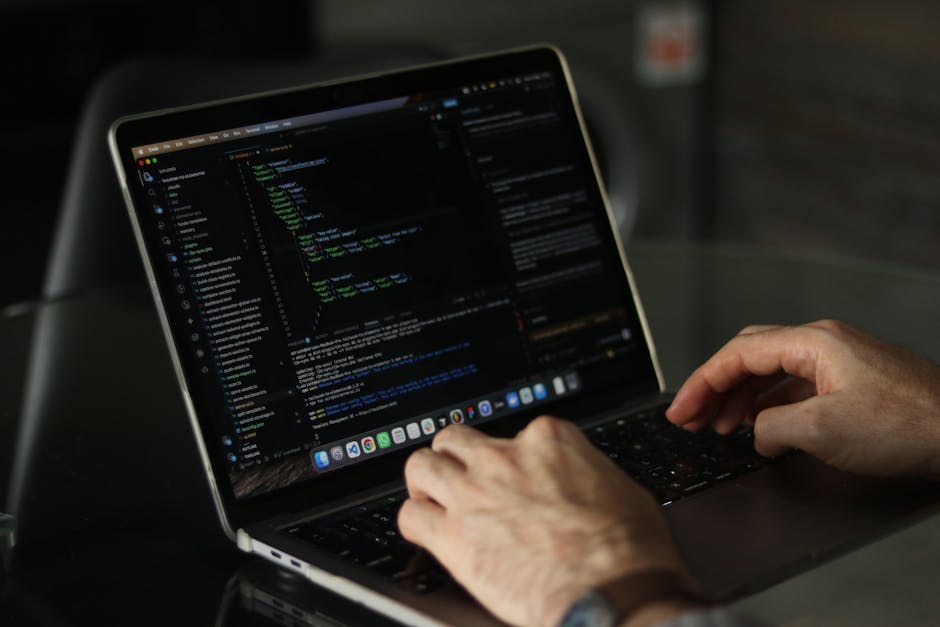 A person operating a silver laptop on a dark surface, with their hands in focus as they type. The laptop screen displays lines of colorful code and text in a programming environment against a dark bac